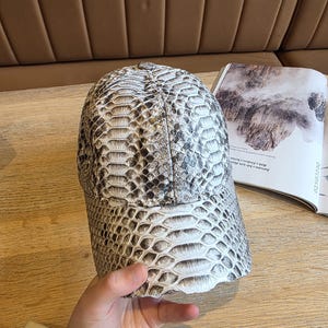 May include: A baseball cap with a snakeskin pattern in shades of gray and white. The cap has a curved brim and a structured crown. The hat is made of a textured material and appears to be leather or a leather-like material. The cap is a fashion accessory.