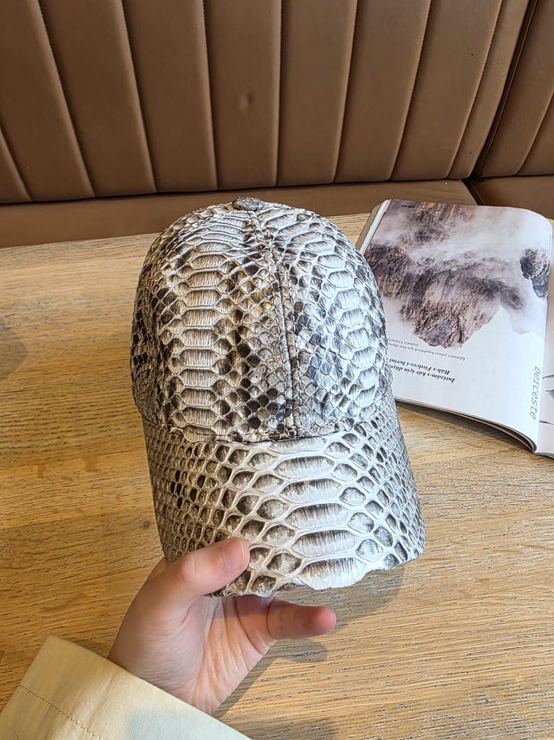 May include: A baseball cap with a snakeskin pattern in shades of gray and white. The cap has a curved brim and a structured crown. The hat is made of a textured material and appears to be leather or a leather-like material. The cap is a fashion accessory.