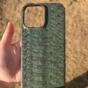 May include: A green phone case with a textured, scaly pattern. The case has a rectangular camera cutout and a dark border. The case is held in a person's hand against a blurred outdoor background.