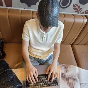 May include: A black cap with a textured pattern is worn by a person sitting at a table. The person is wearing a white polo shirt and blue jeans. A laptop and a magazine are on the table. A black bag is on the table.