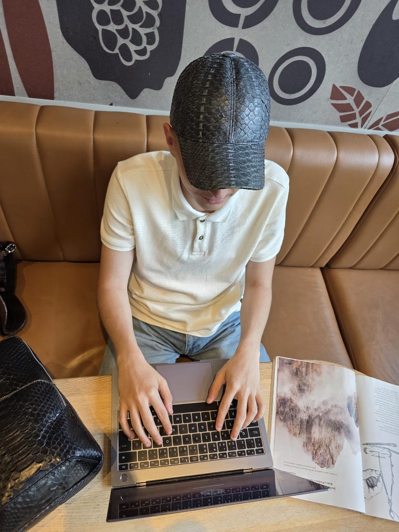 May include: A black cap with a textured pattern is worn by a person sitting at a table. The person is wearing a white polo shirt and blue jeans. A laptop and a magazine are on the table. A black bag is on the table.