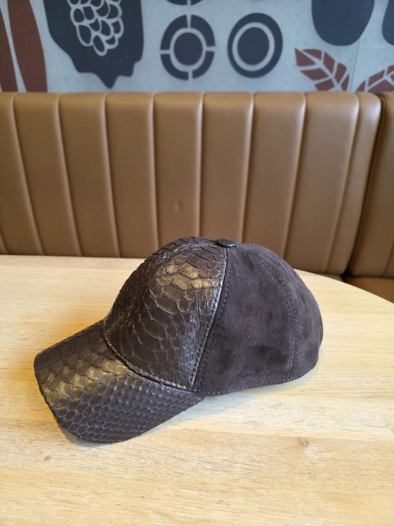 May include: A brown baseball cap with a snakeskin-textured visor and a suede crown. The cap is dark brown and has a button on top. The hat is sitting on a light-colored wooden surface.