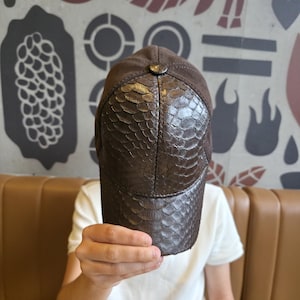 Może przedstawiać: Brązowa czapka bejsbolowa z daszkiem o fakturze skóry węża i zamszową koroną. Czapka ma guzik na górze i zakrzywiony daszek. Kapelusz jest trzymany przed osobą.