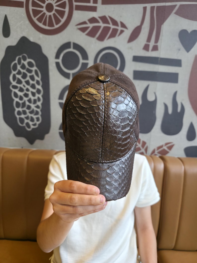Op de afbeelding: Een bruine baseballcap met een vizier met slangenleertextuur en een su&egrave;de kroon. De cap heeft een knoop bovenop en een gebogen klep. De hoed wordt voor een persoon omhoog gehouden.