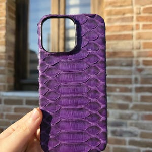 Könnte beinhalten: Eine leuchtend lila Handyhülle mit Schlangenhautstruktur. Die Hülle hat einen Kameraausschnitt und wird vor einer Backsteinmauer gehalten. Die Hülle soll das Telefon vor Kratzern und Beschädigungen schützen.