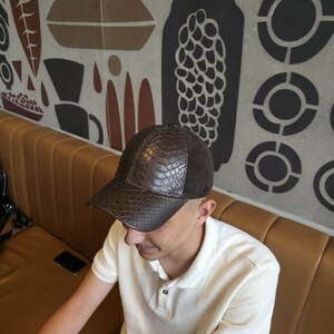 Op de afbeelding: Een bruine baseballcap met een textuurachtig, reptielachtig patroon aan de voorkant en een donkerbruin su&egrave;de bovenstuk. De pet wordt gedragen door een persoon die een witte poloshirt draagt.