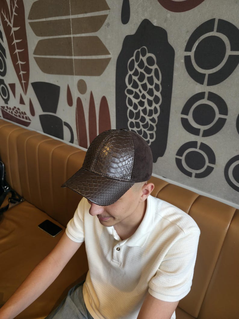 Op de afbeelding: Een bruine baseballcap met een textuurachtig, reptielachtig patroon aan de voorkant en een donkerbruin su&egrave;de bovenstuk. De pet wordt gedragen door een persoon die een witte poloshirt draagt.