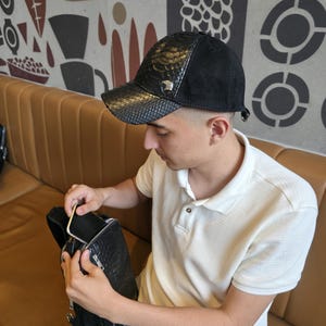 K&ouml;nnte beinhalten: Schwarze Baseballkappe mit Schlangenleder-Struktur und schwarzem Wildleder-Oberteil. Die Kappe hat ein silberfarbenes Emblem. Die Person im Bild tr&auml;gt ein wei&szlig;es Poloshirt und blaue Jeans.