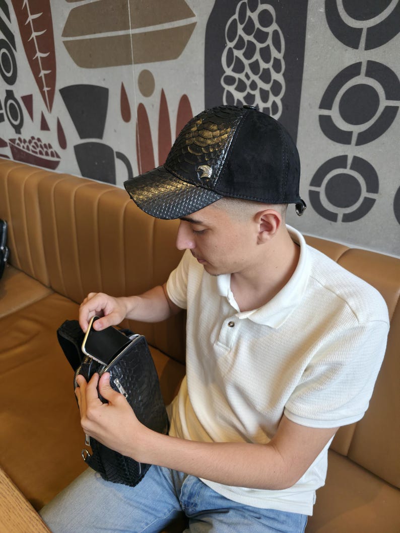 K&ouml;nnte beinhalten: Schwarze Baseballkappe mit Schlangenleder-Struktur und schwarzem Wildleder-Oberteil. Die Kappe hat ein silberfarbenes Emblem. Die Person im Bild tr&auml;gt ein wei&szlig;es Poloshirt und blaue Jeans.