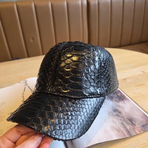 K&ouml;nnte beinhalten: Schwarze Baseballkappe mit einem strukturierten, schuppenartigen Muster. Die Kappe hat einen gebogenen Schirm und liegt auf einem offenen Buch. Die Kappe scheint aus Leder oder einem &auml;hnlichen Material zu sein.