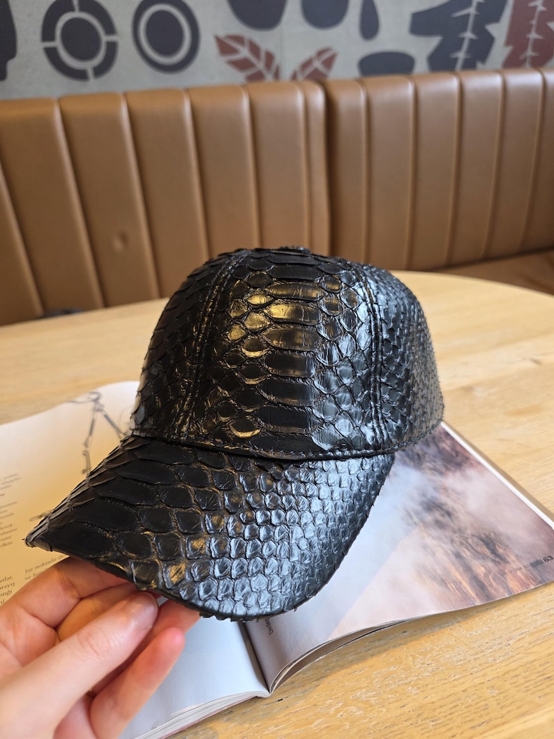 K&ouml;nnte beinhalten: Schwarze Baseballkappe mit einem strukturierten, schuppenartigen Muster. Die Kappe hat einen gebogenen Schirm und liegt auf einem offenen Buch. Die Kappe scheint aus Leder oder einem &auml;hnlichen Material zu sein.