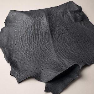 May include: A sheet of dark grey leather with a textured surface. The leather features a pattern of small, evenly spaced bumps. The edges are uneven, indicating it is a hide. The overall appearance is of a high-quality material.
