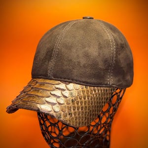 May include: A brown baseball cap with a textured, suede-like crown and a gold, snakeskin-patterned brim. The cap is displayed on a black, wire-frame head form against an orange background.