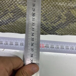 Peut inclure: Gros plan sur un tissu &agrave; motifs vert olive et noir. Une r&egrave;gle m&eacute;tallique indique les mesures en centim&egrave;tres. Le tissu pr&eacute;sente un motif textur&eacute; en forme d'&eacute;cailles. La r&egrave;gle mesure jusqu'&agrave; 43 cm.