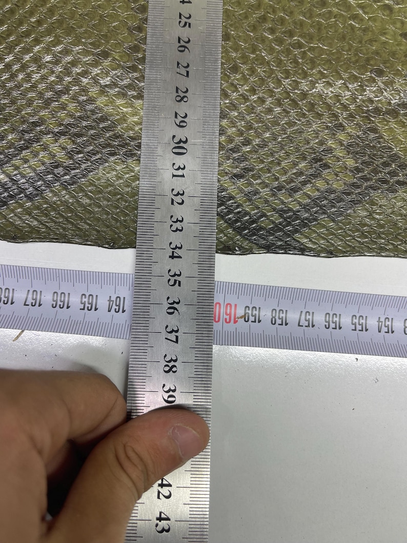Peut inclure: Gros plan sur un tissu &agrave; motifs vert olive et noir. Une r&egrave;gle m&eacute;tallique indique les mesures en centim&egrave;tres. Le tissu pr&eacute;sente un motif textur&eacute; en forme d'&eacute;cailles. La r&egrave;gle mesure jusqu'&agrave; 43 cm.