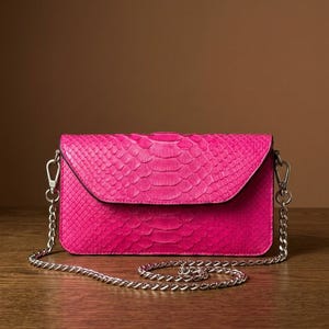 May include: A rectangular hot pink clutch bag featuring a textured snakeskin pattern. The bag has a fold-over flap closure and is equipped with a detachable silver-toned metal chain shoulder strap that attaches to the sides with lobster claw clasps.