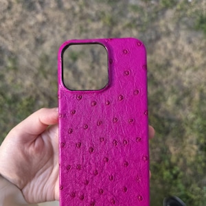 Peut inclure: Un étui de téléphone magenta avec un motif texturé et pointillé. L'étui a une forme rectangulaire avec une découpe pour l'appareil photo. L'étui est tenu dans la main d'une personne sur un arrière-plan extérieur flou.
