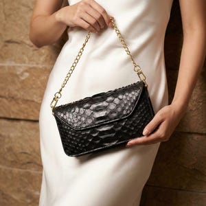 Pode incluir: Clutch preta com um padrão texturizado semelhante à pele de réptil. A bolsa tem uma alça de corrente dourada e um fecho de aba. A clutch é segurada por uma pessoa que usa um vestido branco.