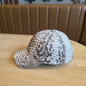 May include: A baseball cap with a snakeskin pattern in shades of gray, white, and black. The cap is sitting on a wooden table. The hat has a curved brim and a structured crown.