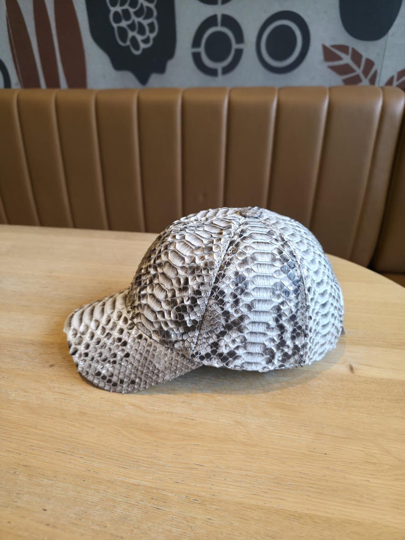 May include: A baseball cap with a snakeskin pattern in shades of gray, white, and black. The cap is sitting on a wooden table. The hat has a curved brim and a structured crown.