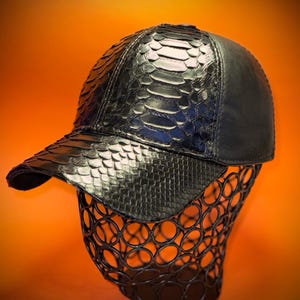 May include: Black baseball cap with a snakeskin pattern on the front and bill. The cap is displayed on a black wire form against an orange background. The cap has a curved bill and a structured crown.