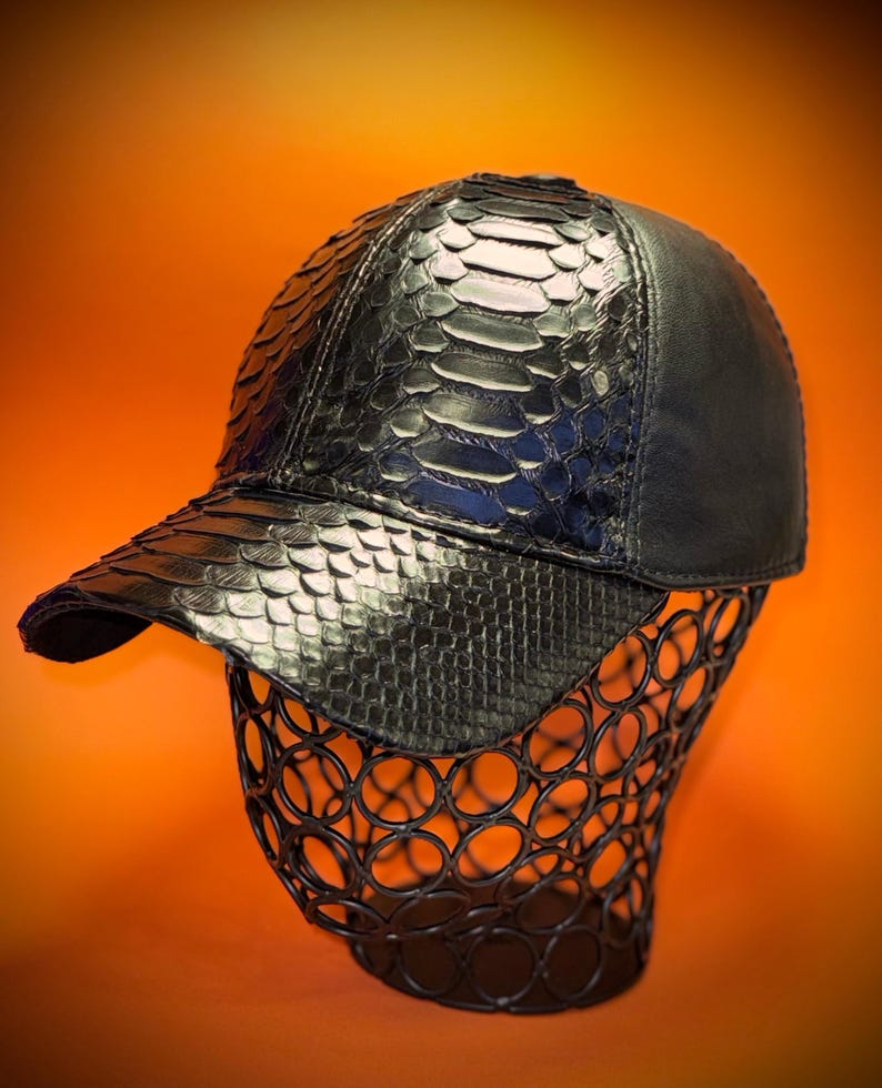 May include: Black baseball cap with a snakeskin pattern on the front and bill. The cap is displayed on a black wire form against an orange background. The cap has a curved bill and a structured crown.