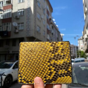May include: A yellow and black snakeskin-patterned wallet with a black border. The rectangular wallet is made of leather. The wallet is held up in front of a blurred background of buildings and a blue sky.
