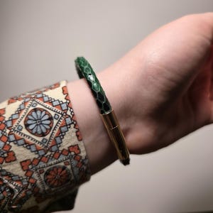 以下が含まれることがあります： 金色の留め金が付いた、緑と黒の模様のブレスレットが手首に表示されています。 ブレスレットは、ヘビのような質感があります。 柄物のシャツの袖が背景に見えます。