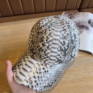 May include: A baseball cap with a snakeskin pattern in shades of gray and white. The cap has a curved brim and a structured crown, offering a stylish accessory. The hat is made of a textured material.