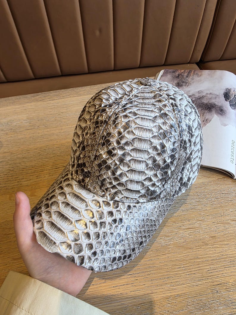 May include: A baseball cap with a snakeskin pattern in shades of gray and white. The cap has a curved brim and a structured crown, offering a stylish accessory. The hat is made of a textured material.