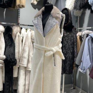 Peut inclure: Un gilet de fourrure sans manches crème avec une ceinture assortie est présenté sur un mannequin. Le gilet a un col et un revers à motif peau de serpent. D'autres manteaux et vestes en fourrure sont suspendus en arrière-plan.