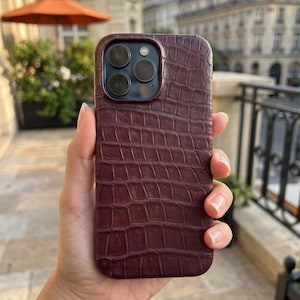 May include: A dark burgundy smartphone case featuring a textured crocodile skin pattern. The case has a precise cutout for the triple-lens camera system and flash, covering the back of the device while maintaining access to the camera module.