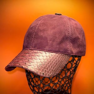 Peut inclure: Casquette de baseball bordeaux avec une visière à motif peau de serpent. La casquette est présentée sur un support en fil de fer noir sur fond orange. La couronne de la casquette a des coutures visibles.