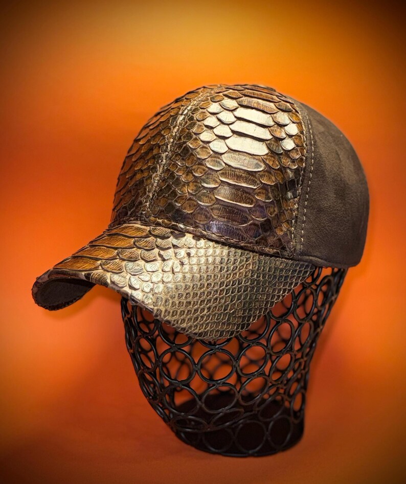 May include: A baseball cap with a snakeskin pattern on the front and brim, and a brown suede back. The cap is displayed on a black wire form against an orange background.