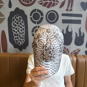 May include: A baseball cap with a snakeskin pattern in shades of white, grey, and brown. The cap is held up in front of a person. The background features a wall with various brown and grey shapes.
