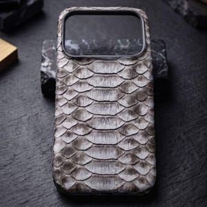 May include: A rectangular smartphone case featuring a textured, grey and white snakeskin pattern. The case has a black frame surrounding the camera cutout at the top and a smooth, consistent scale design covering the entire back surface.