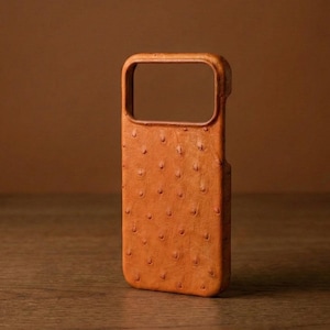 Puede incluir: Funda de teléfono de color marrón anaranjado con un recorte rectangular para la cámara. La funda tiene una superficie texturizada y con bultos. Está hecha de un material similar al cuero y está diseñada para proteger un smartphone.