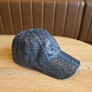 May include: A dark gray baseball cap with a textured, scaly pattern. The cap has a curved brim and is made of a material that resembles snakeskin. The cap is sitting on a light brown wooden surface.