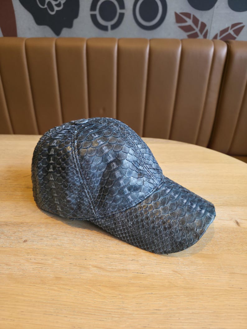 May include: A dark gray baseball cap with a textured, scaly pattern. The cap has a curved brim and is made of a material that resembles snakeskin. The cap is sitting on a light brown wooden surface.