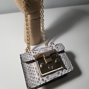 May include: A miniature handbag with a gold chain strap. The bag has a white and grey snakeskin pattern and a gold clasp. The bag is approximately 10 cm wide and 7.5 cm tall.