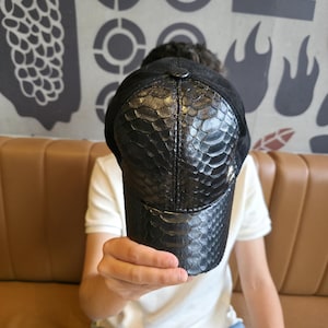 K&ouml;nnte beinhalten: Schwarze Baseballkappe mit Schlangenmuster auf der Vorderseite und einem schwarzen Wildleder-Oberteil. Die Kappe wird vor einer Person hochgehalten. Der Hintergrund zeigt eine braune Lederbank und eine Wand mit abstrakten Designs.