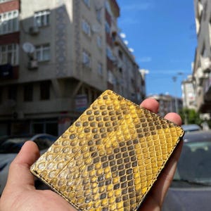 May include: A rectangular wallet with a yellow and black snakeskin pattern. The wallet is made of leather and is held in a hand. The pattern consists of yellow and black diamond shapes. The background shows buildings and a blue sky.