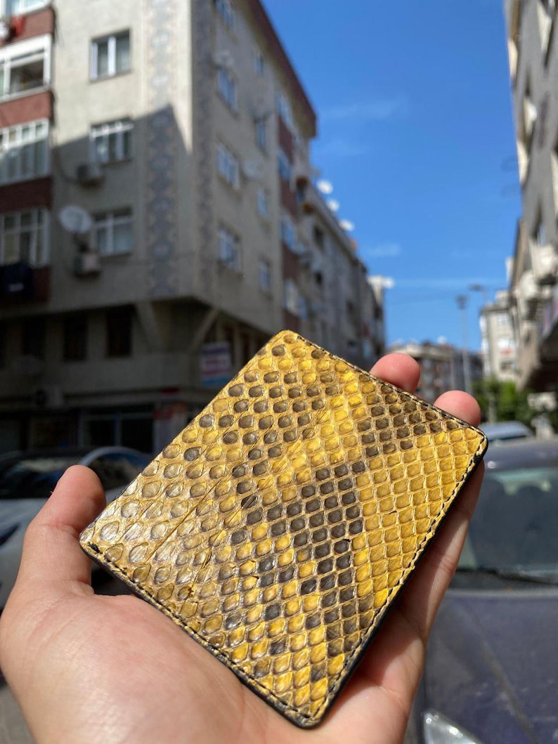 May include: A rectangular wallet with a yellow and black snakeskin pattern. The wallet is made of leather and is held in a hand. The pattern consists of yellow and black diamond shapes. The background shows buildings and a blue sky.