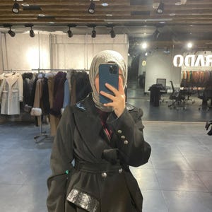 Könnte beinhalten: Ein schwarzer Mantel mit Wickeldesign, Gürtel in der Taille und dekorativen Knöpfen. Der Mantel hat ein einzigartiges Taschen Detail mit einem strukturierten, glänzenden Akzent. Der Mantel wird in einem Bekleidungsgeschäft präsentiert.