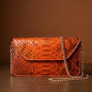 May include: A rectangular orange handbag featuring a textured snakeskin pattern across the exterior. The bag includes a fold-over flap closure and a thin, metallic gold-toned chain shoulder strap attached to the sides with small lobster clasps.