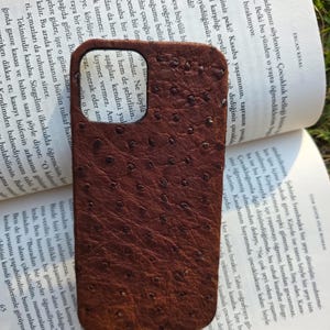 Pode incluir: Uma capa de telefone de couro marrom com uma superf&iacute;cie texturizada, repousando sobre um livro aberto. A capa tem uma cor escura e rica e um padr&atilde;o de pequenos pontos espa&ccedil;ados.