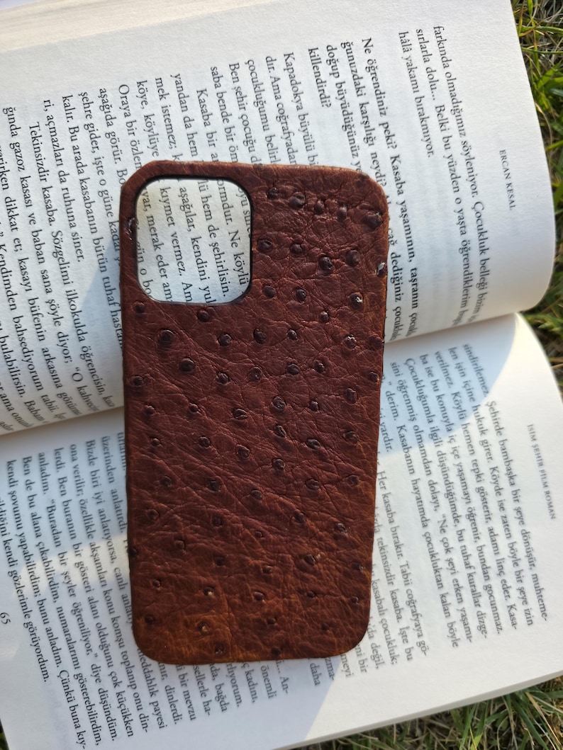 Pode incluir: Uma capa de telefone de couro marrom com uma superf&iacute;cie texturizada, repousando sobre um livro aberto. A capa tem uma cor escura e rica e um padr&atilde;o de pequenos pontos espa&ccedil;ados.