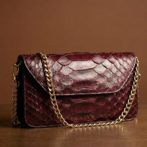 May include: A burgundy clutch bag with a gold chain strap. The bag has a snakeskin-like texture and a flap closure. The background is a solid brown colour.
