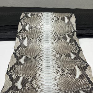 Może przedstawiać: Kawałek skóry węża z wzorem szarych, białych i brązowych łusek. Skóra węża jest złożona, odsłaniając czarny materiał skórzany w tle. Skóra węża ma teksturowaną powierzchnię.