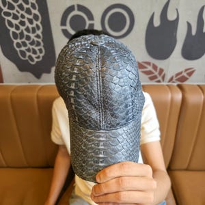 Może przedstawiać: Ciemnoszara czapka bejsbolowa z teksturowanym, łuskowatym wzorem. Czapka ma zakrzywiony daszek i strukturalną koronę. Materiał wydaje się być skórą lub materiałem skóropodobnym. Czapka jest trzymana przed osobą.